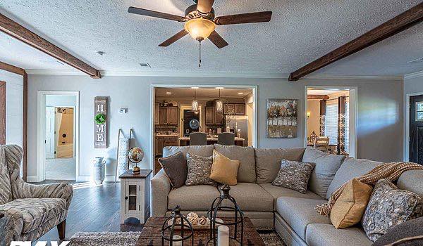 ```plaintext
A cozy living room with a large beige sectional sofa adorned with patterned pillows, a wooden coffee table, and a ceiling fan. The room opens to a warmly lit kitchen.
```