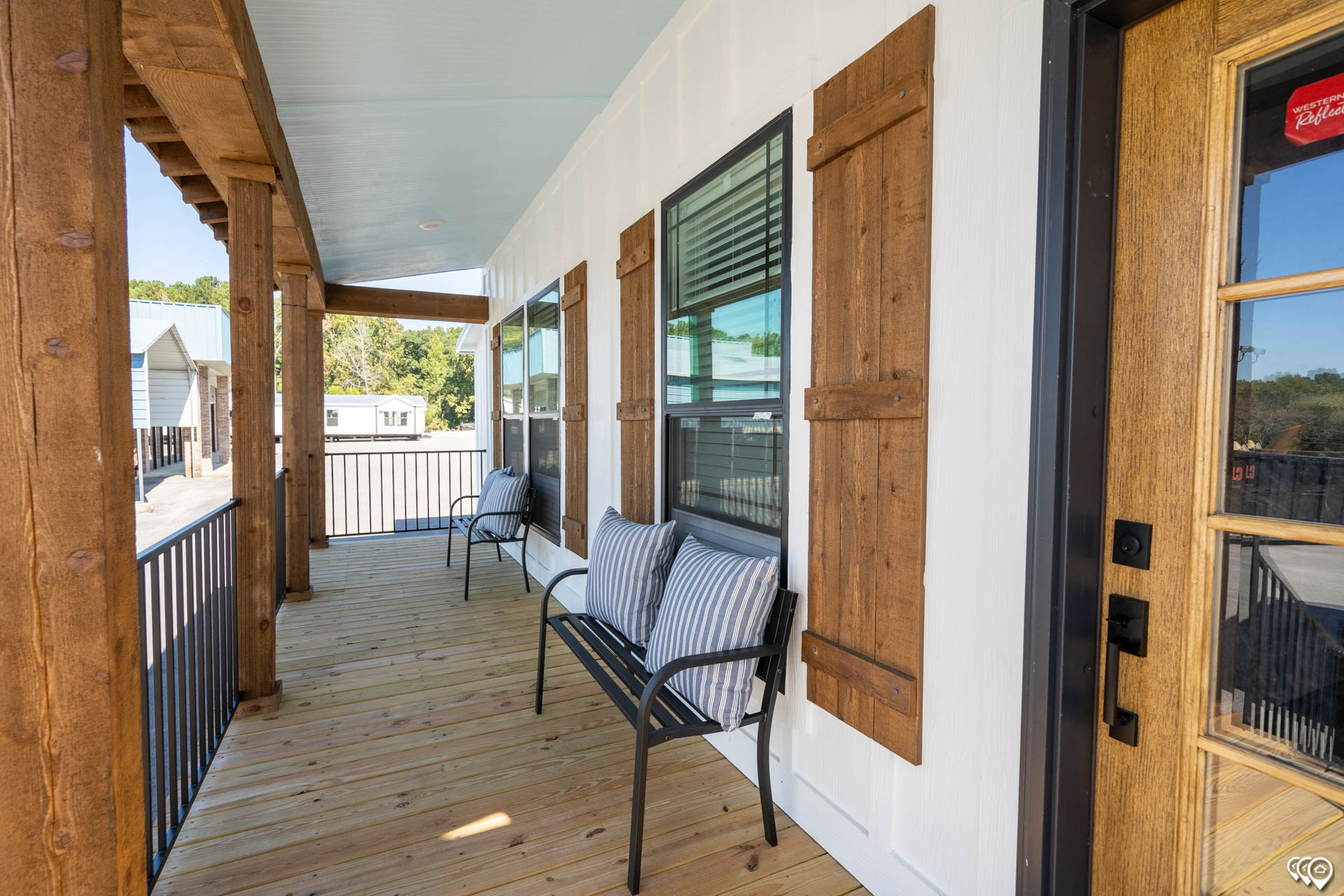A wooden porch features two benches with striped cushions, framed by rustic shutters and railing. Sunlight casts soft shadows, creating a relaxed atmosphere.