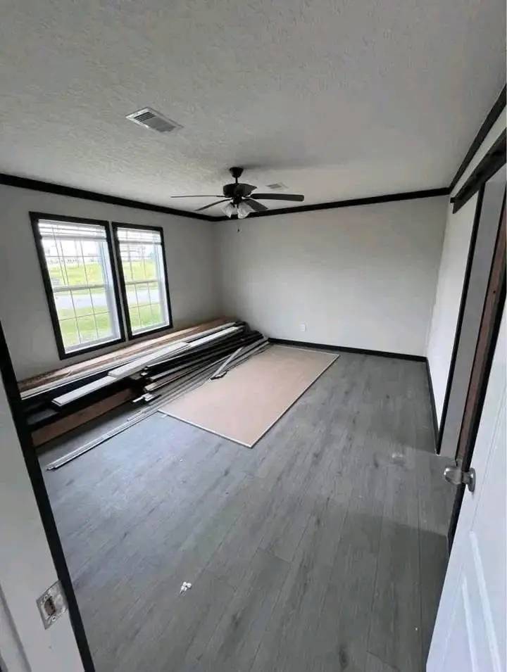 A sparsely furnished room features gray wood flooring, two large windows, a ceiling fan, and construction materials on the floor, suggesting renovation.