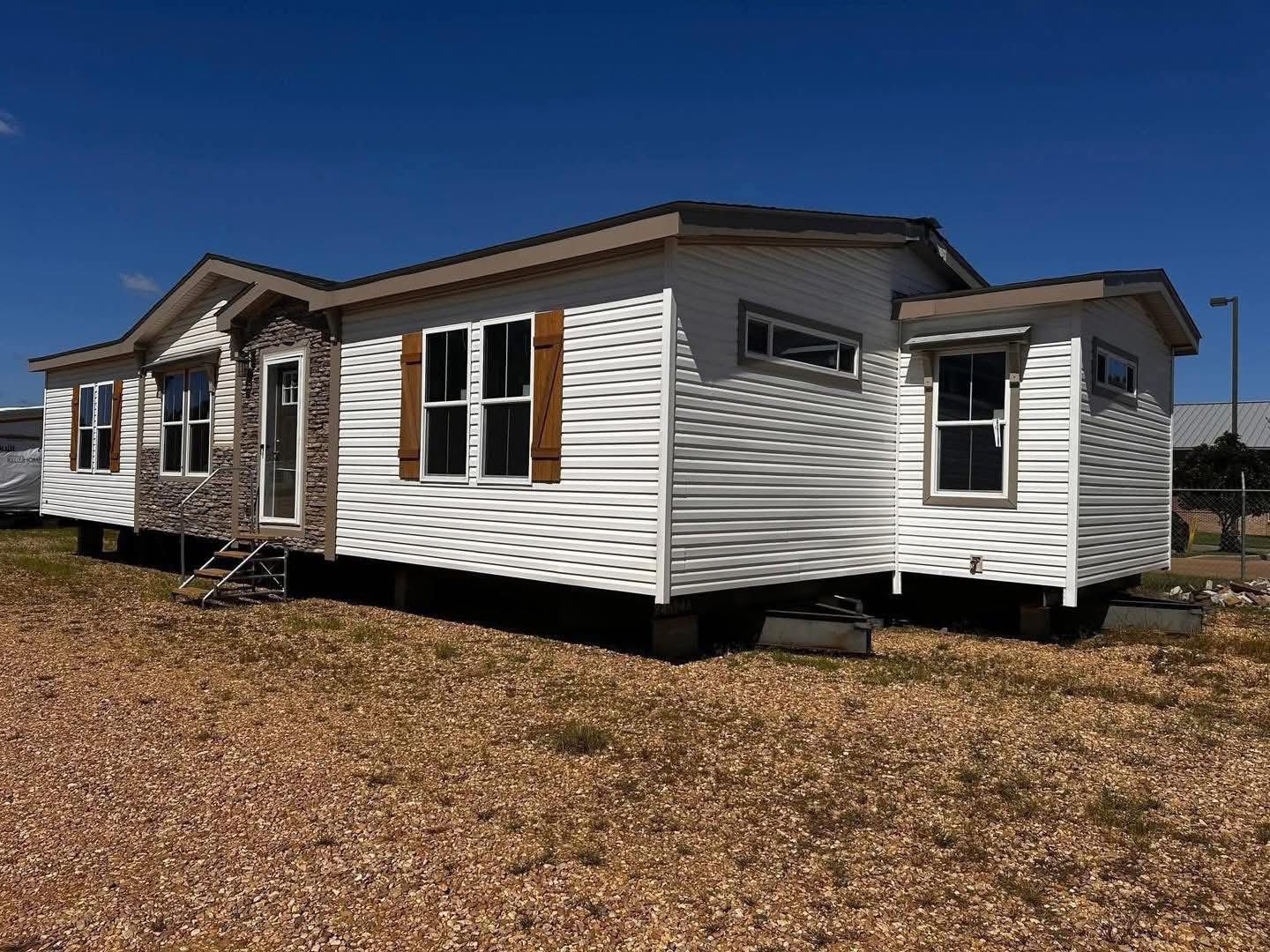 A white modular home with brown shutters and a stone accent is set on gravel. A small metal staircase leads to the door. The sky is clear and blue.