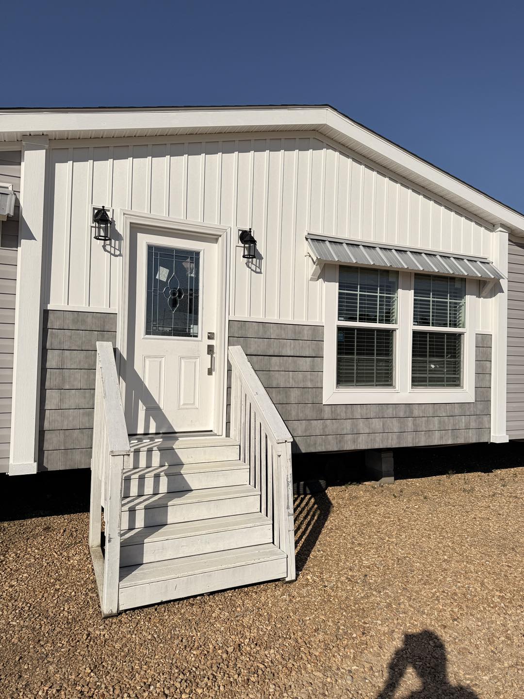 Small white and gray modular home with a decorative glass door and two windows. Simple porch with railings, set under a clear blue sky.