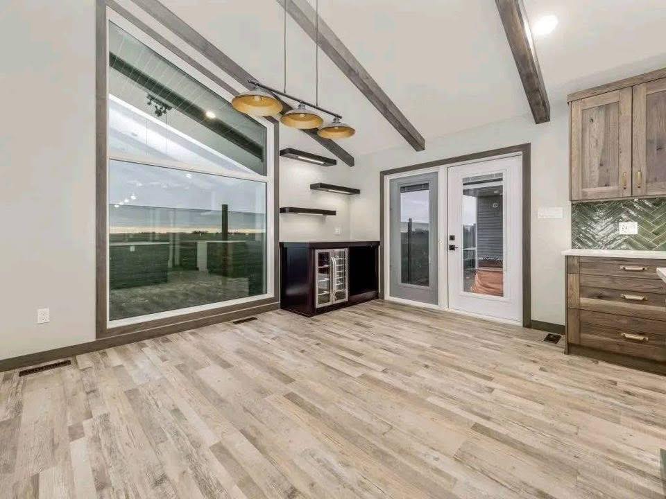 Modern room with wood flooring, large windows, and exposed beams. Featuring a wine fridge, floating shelves, and pendant lights, it conveys a sleek, airy vibe.
