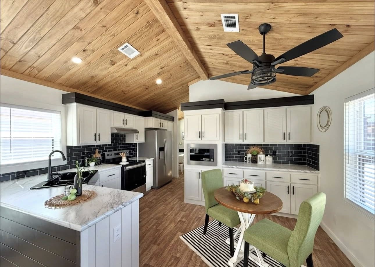 Spacious kitchen with wooden ceiling and floors, modern appliances, black and white cabinets, green chairs, round table, and ceiling fan for a cozy atmosphere.