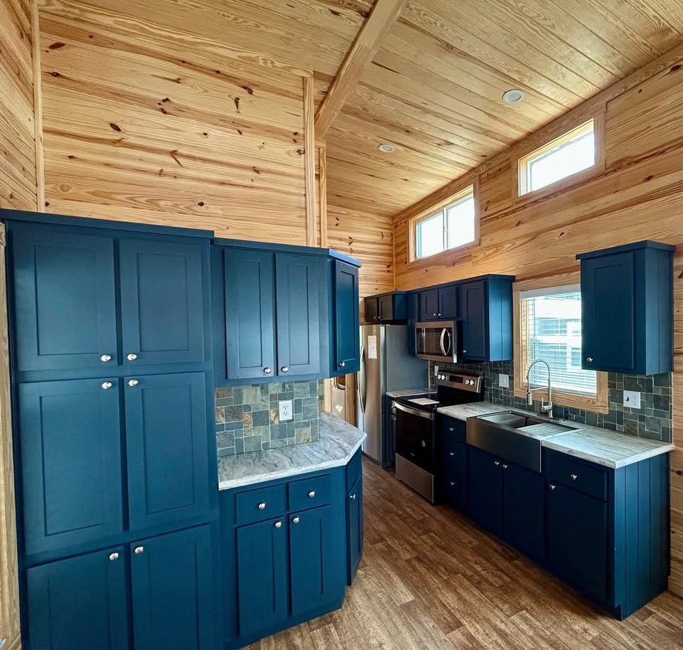 Rustic kitchen with blue cabinets, wooden walls, and stone backsplash. Features stainless steel appliances and abundant natural light from high windows.
