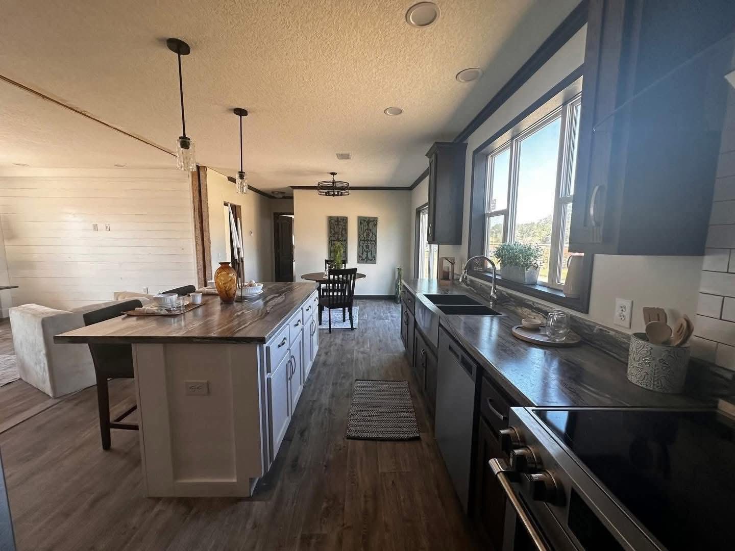 Modern kitchen with island, pendant lights, and wood floor. A window above the sink lets in natural light. Dining area visible in the background.