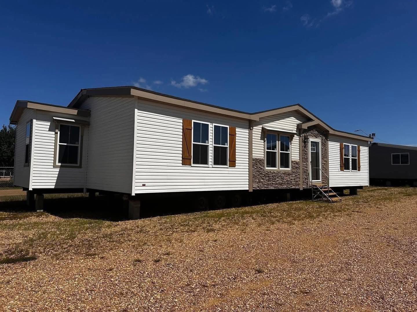 A single-story modular home with white siding, brown shutters, and a stone facade sits on a gravel lot under a clear blue sky, conveying a rural, serene setting.
