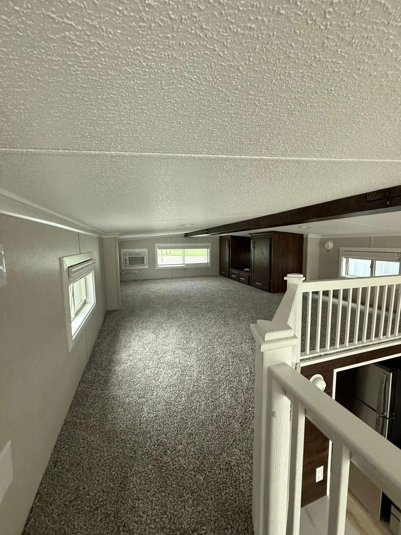 Cozy loft space with gray carpet, white railing, and a low ceiling. Windows provide natural light, while dark wood cabinets add warmth. Airy and inviting.