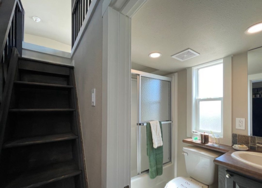 A small, bright bathroom with a shower stall, frosted glass doors, and a green towel. Staircase on the left, natural light through a window. Cozy ambiance.