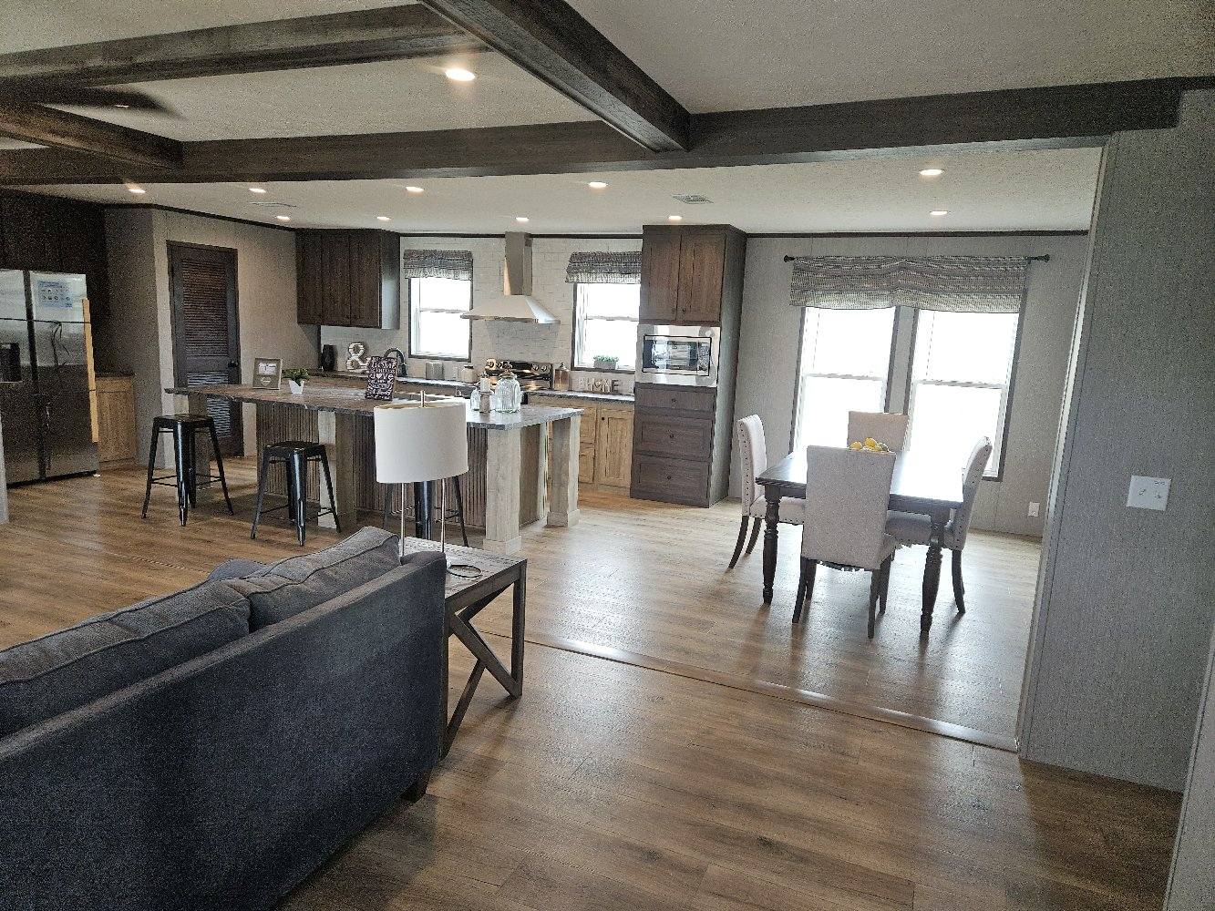 Open-concept kitchen and dining area with wooden floors and ceiling beams. A gray sofa faces a rustic kitchen island with stools. Natural light fills the space, creating a cozy atmosphere.
