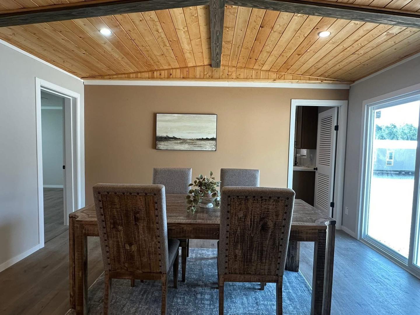 Dining room with rustic wooden table and four chairs, under a wooden ceiling. A landscape painting and plant centerpiece add charm. Bright and cozy.