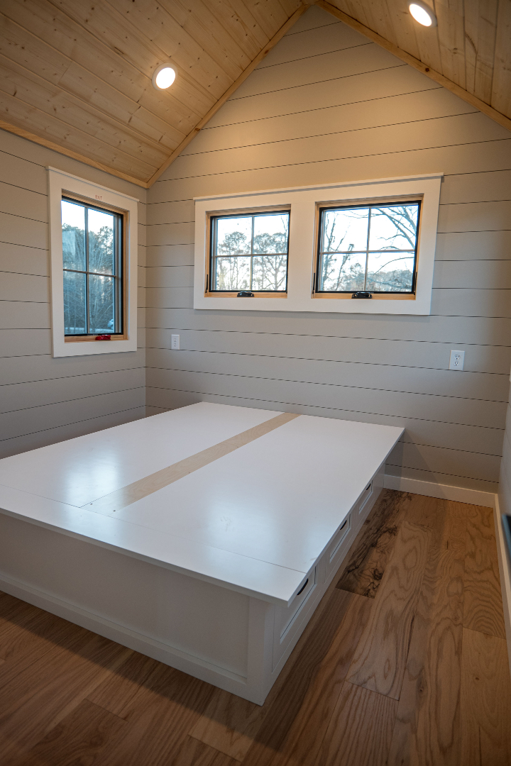 A cozy, minimalist bedroom with light wood walls and ceiling. A large white built-in bed and three small windows fill the space, creating a serene, airy atmosphere.