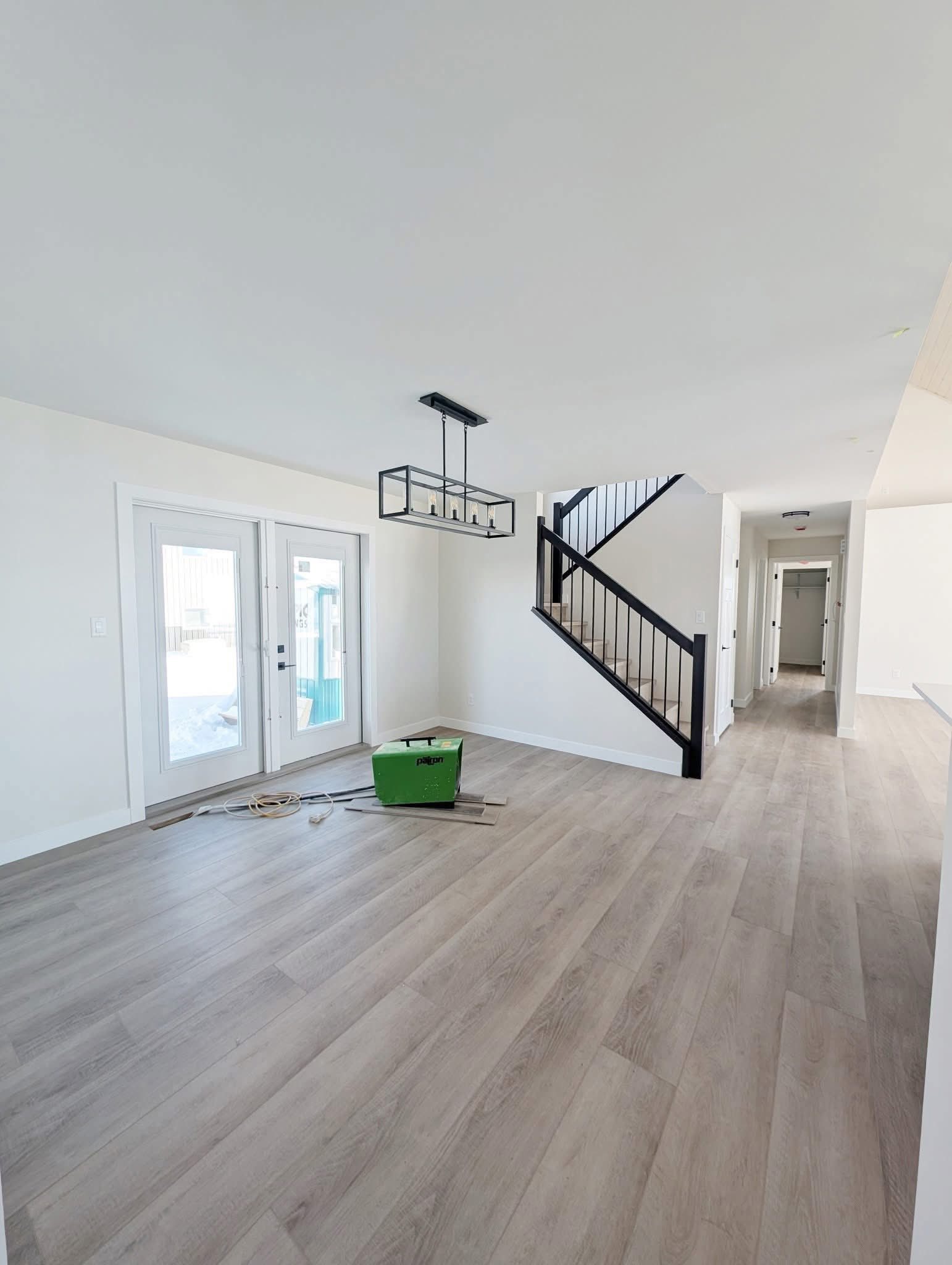 Spacious, bright room with light wood flooring, white walls, and a modern black metal staircase. A large windowed door and tools rest on the floor.