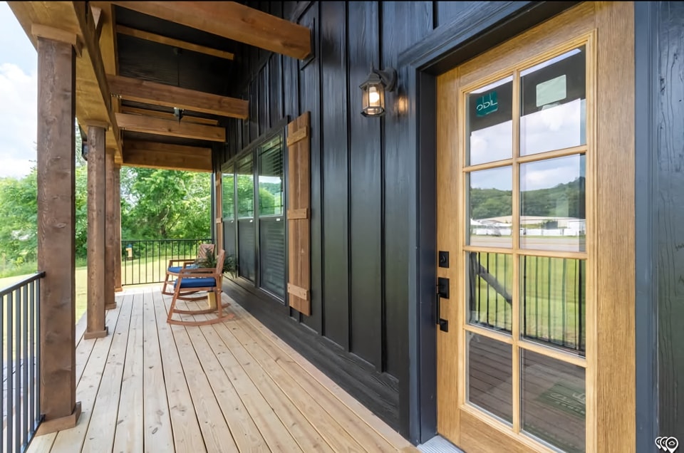 A rustic porch with wooden beams, a tan wooden floor, black siding, a wooden door with glass panels, and a rocking chair creating a cozy, inviting atmosphere.