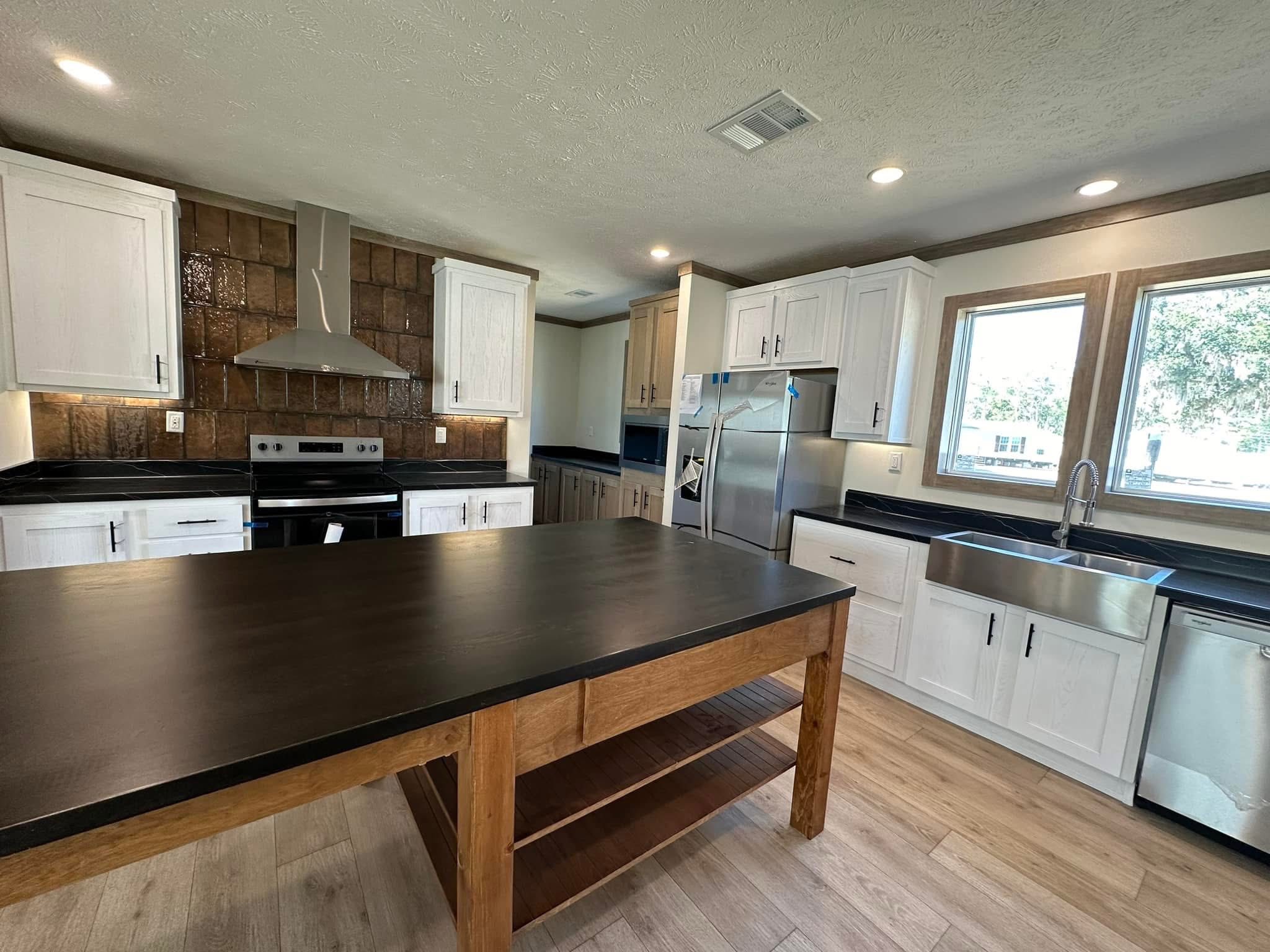 A modern kitchen with wooden floors, a large dark wood island, white cabinets, stainless steel appliances, and ample natural light from two windows.