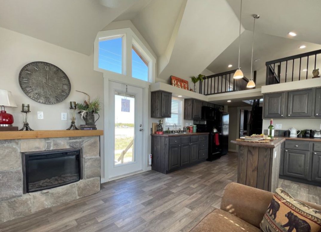 Spacious modern kitchen with dark wood cabinets, high ceilings, and loft. Features a stone fireplace, decorative clock, and large windows. Cozy, inviting atmosphere.