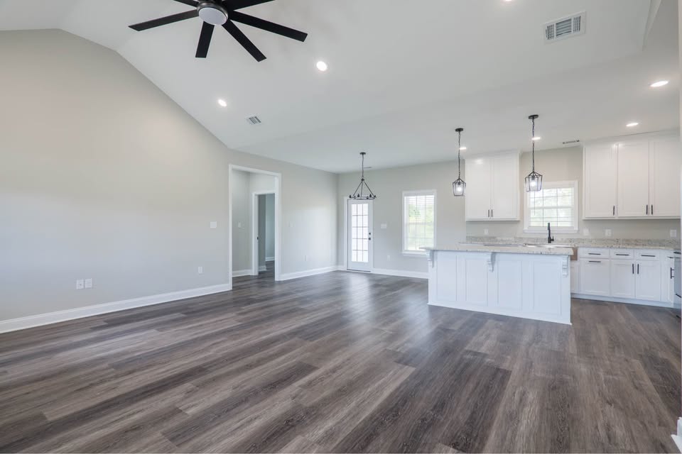 Spacious modern living area with light gray walls and dark wood flooring. Features a sleek ceiling fan, open kitchen with white cabinets, and pendant lights.