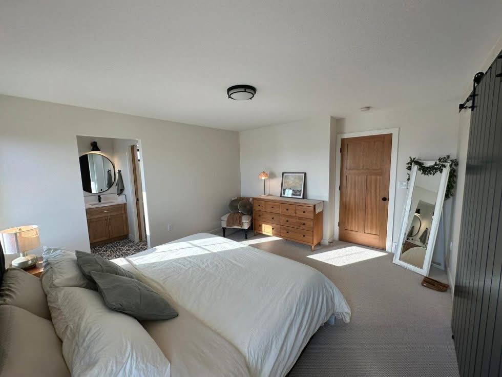 Bright minimalist bedroom with a cozy bed, white linens, wooden dresser, and mirror. A doorway leads to a bathroom with a round mirror. Calm ambiance.