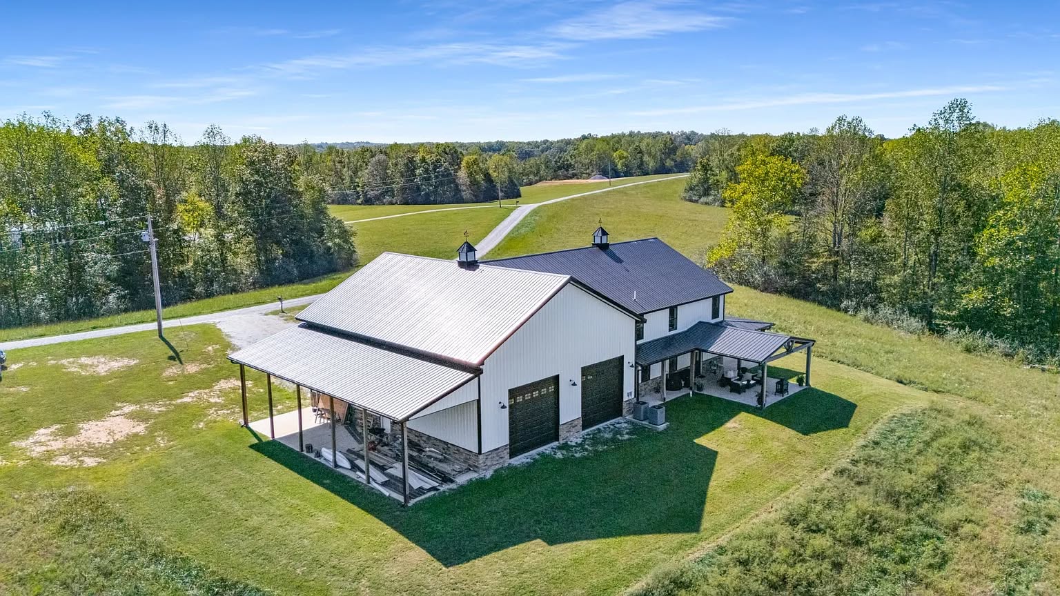 Aerial view of a white barn-style house with a metal roof, surrounded by lush green fields and trees under a clear blue sky, conveying tranquility.
