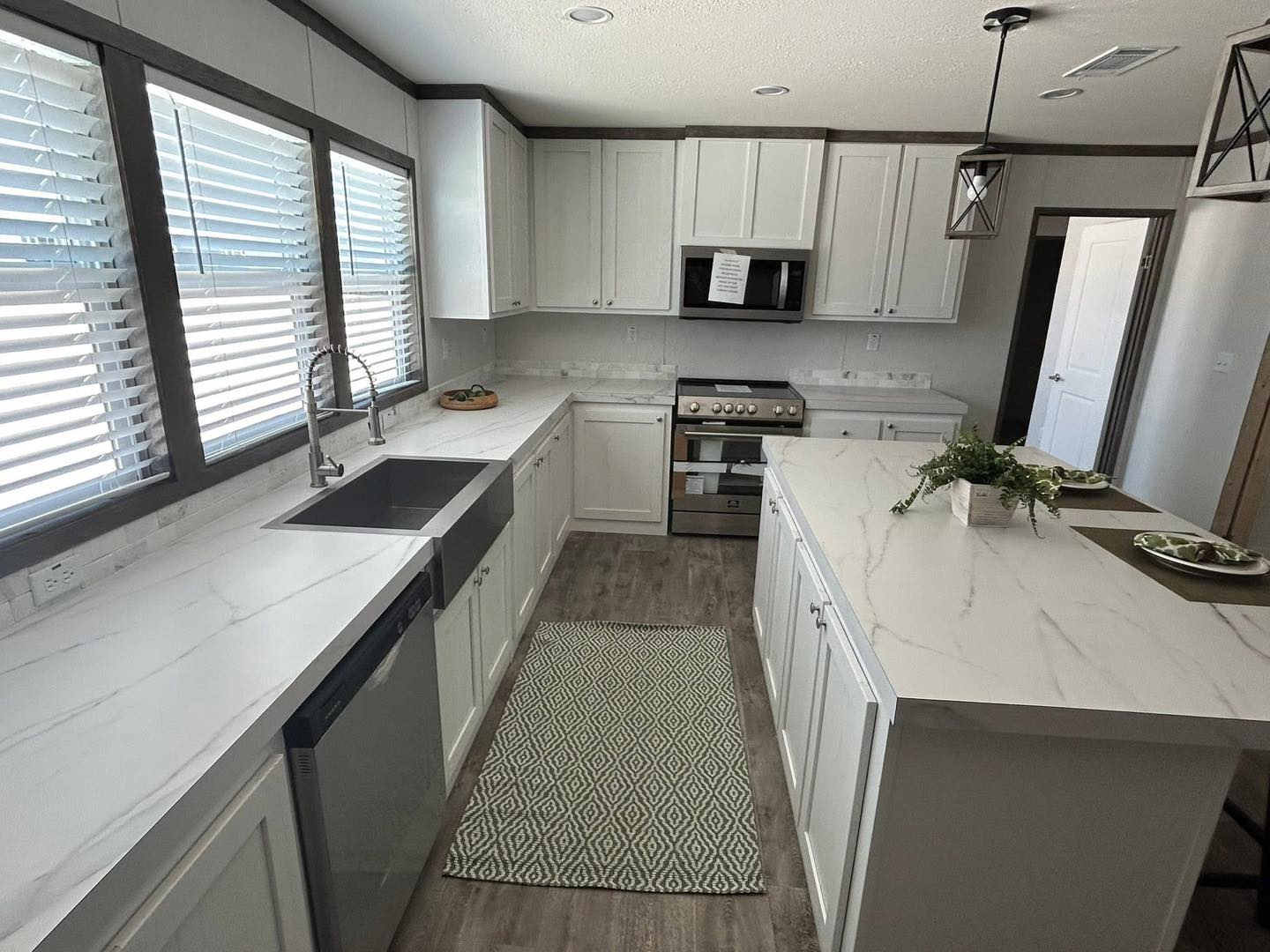 Bright modern kitchen with white cabinets, marble countertops, and stainless steel appliances. Sunlight streams through blinds, creating an airy atmosphere.