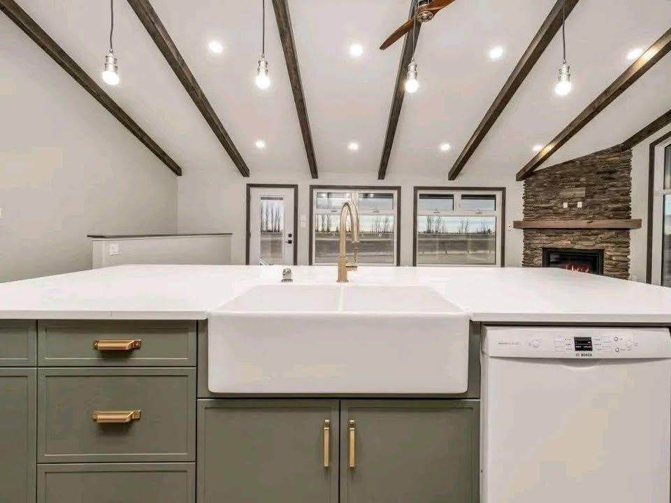 A modern kitchen with white countertops, a large farmhouse sink, and brass fixtures. Vaulted ceiling with wooden beams and hanging lights. Stone fireplace in the background.