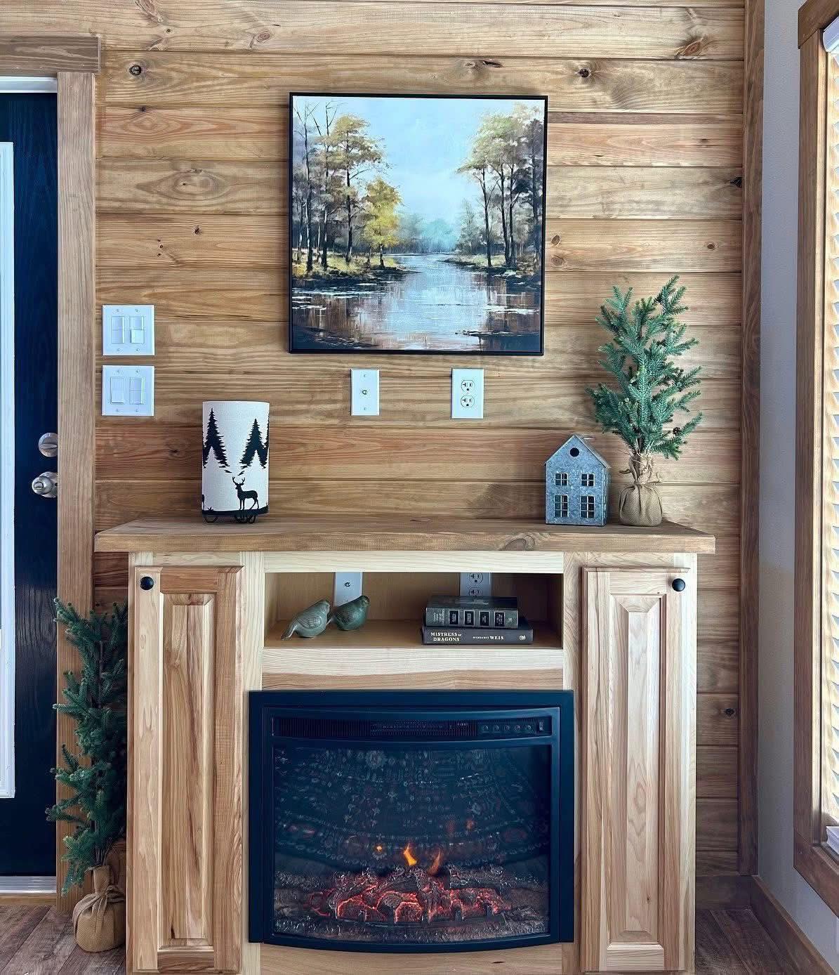 Cozy wooden interior features an electric fireplace, landscape painting, small potted tree, decorative house, and forest-themed candle. Warm, inviting vibe.