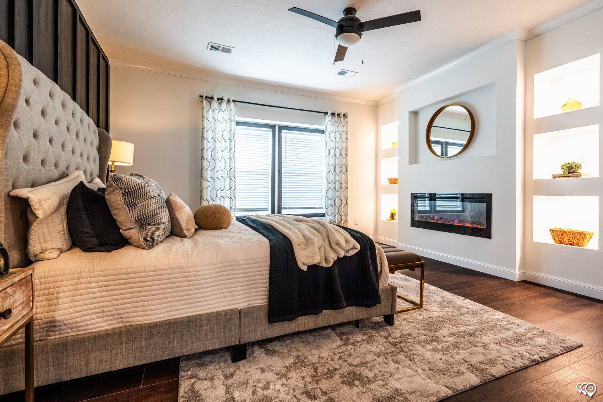 Cozy bedroom with a tufted bed, layered cushions, and a throw. Soft lighting from a fireplace and accent shelves. Elegant, warm, and inviting.