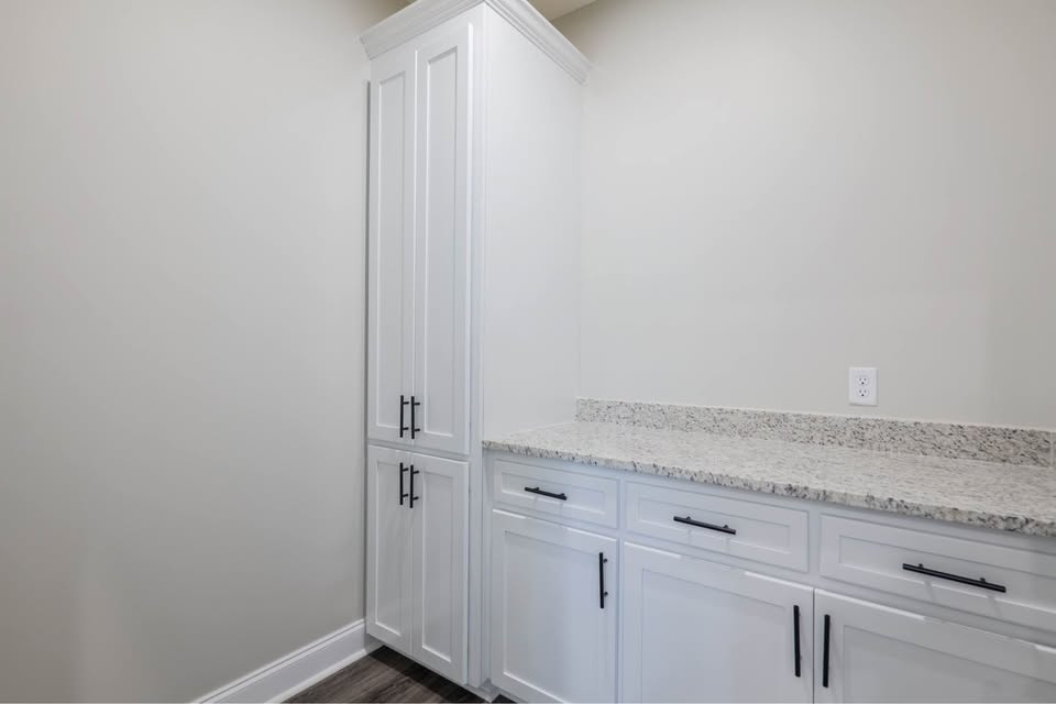 A corner kitchen area with white cabinetry, black handles, and a gray speckled countertop. The space feels modern and minimalistic with a neutral tone.
