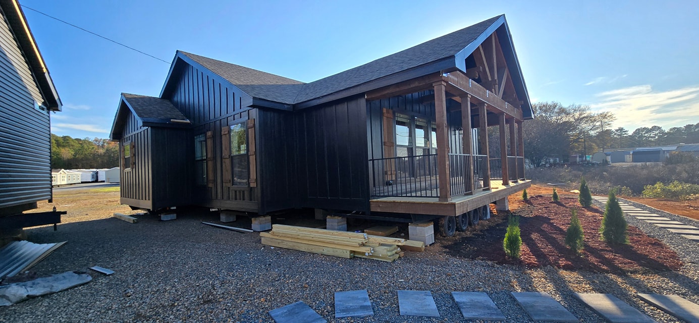 Modern black wooden house elevated on a gravel plot with a surrounding garden. A clear blue sky and distant trees provide a peaceful backdrop.