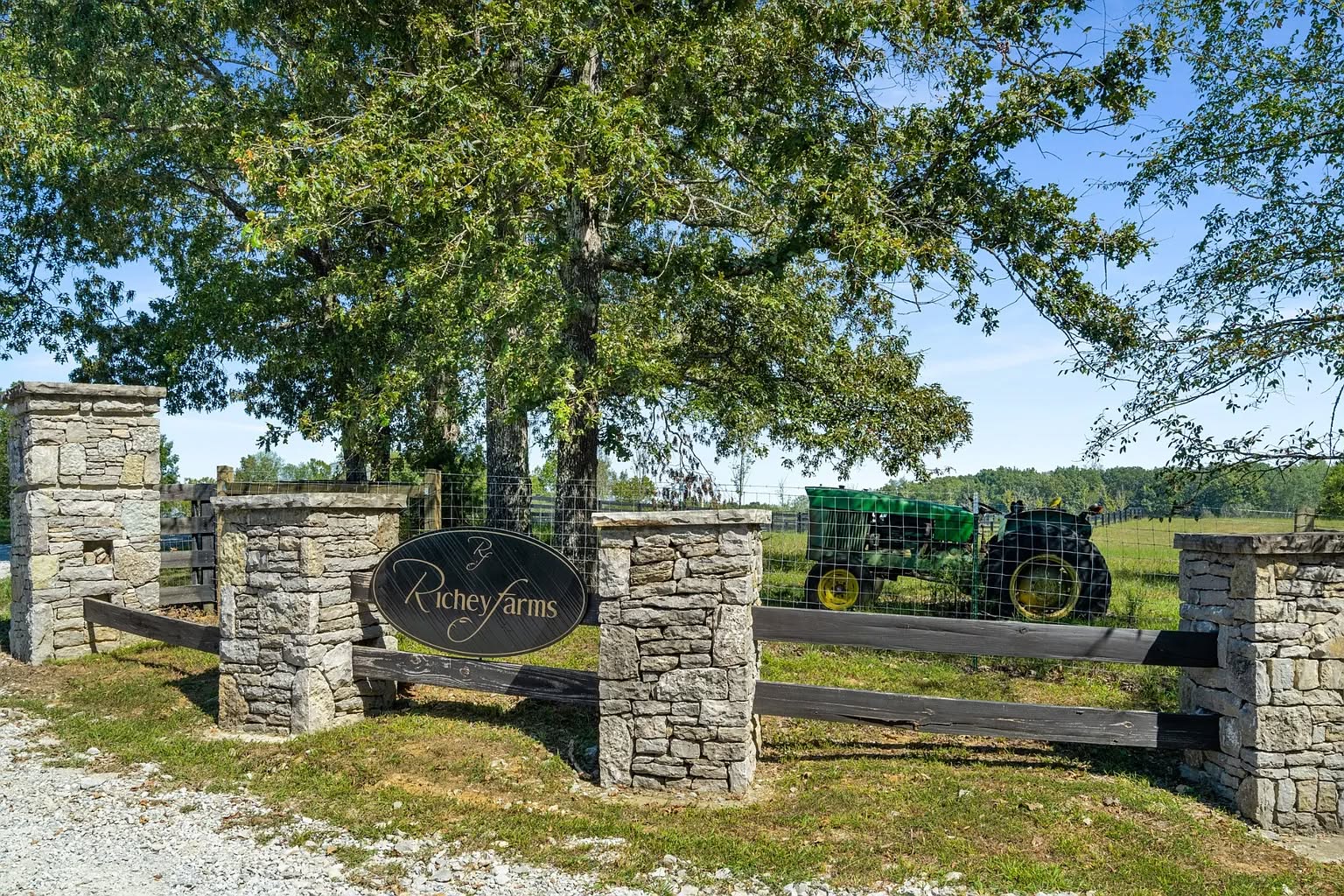 A rustic stone entrance with "Richey Farms" sign leads to a grassy field. A green tractor sits behind the fence. Sunny day with lush trees.