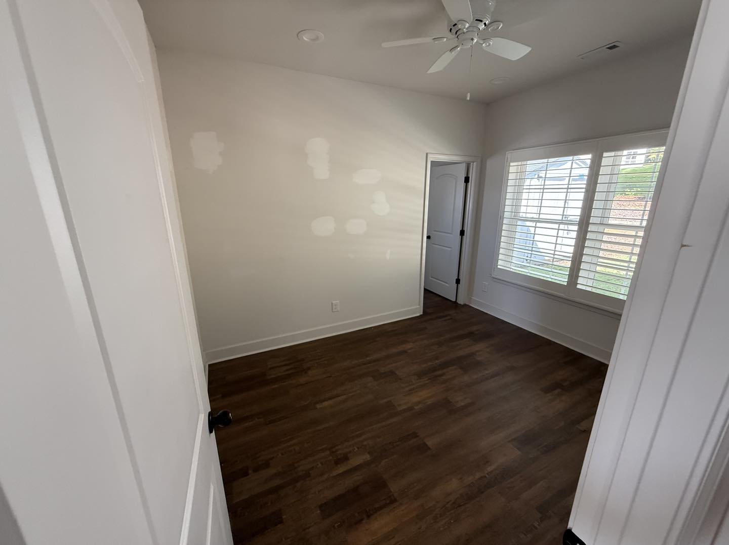 Empty room with hardwood floors, white walls, a ceiling fan, and a large window with blinds. Patches on walls suggest recent repair work. Calm atmosphere.