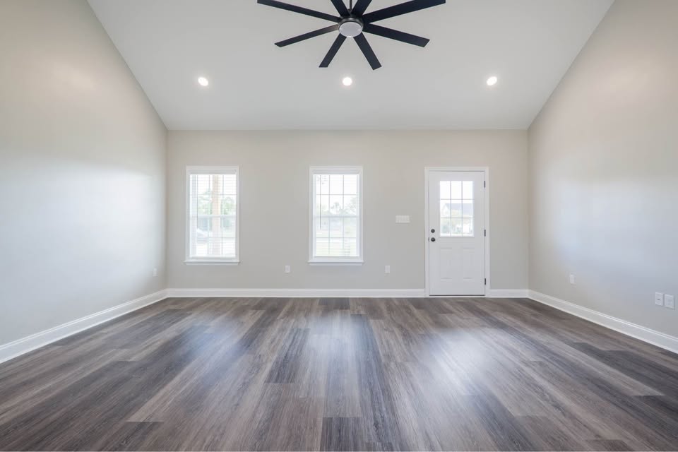 Spacious empty room with light gray walls and a dark wood floor. Features include a modern black ceiling fan, two windows, and a white door, creating an airy atmosphere.