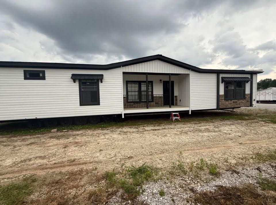 A white manufactured home under a cloudy sky. The front porch has a small step ladder, dark window trim, and brown stone accents, set on a gravel lot.