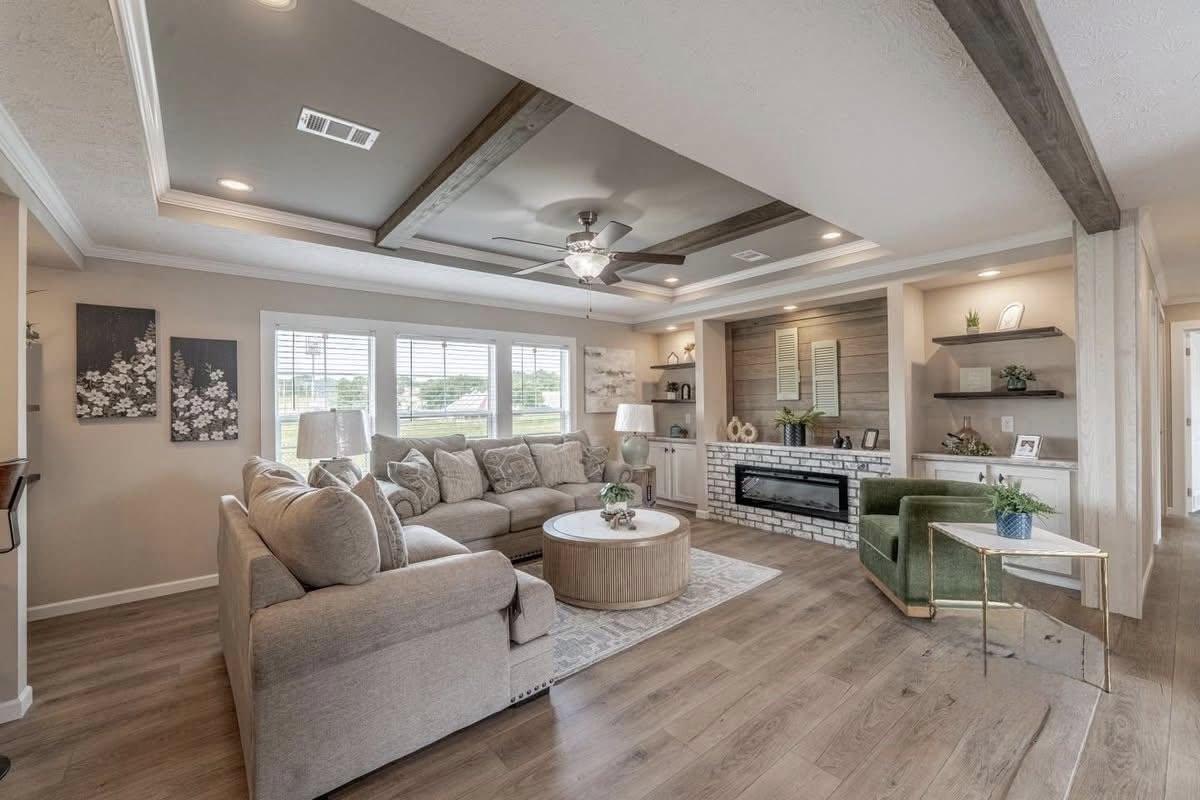 Spacious, cozy living room with beige sectional sofa, round coffee table, green velvet chair, and fireplace. Decor includes plants, art, and soft lighting.