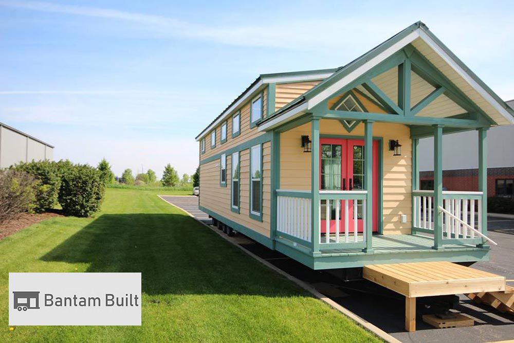 Small beige and teal tiny house with a porch and red door, set on green grass under a clear sky. "Bantam Built" logo in the corner.