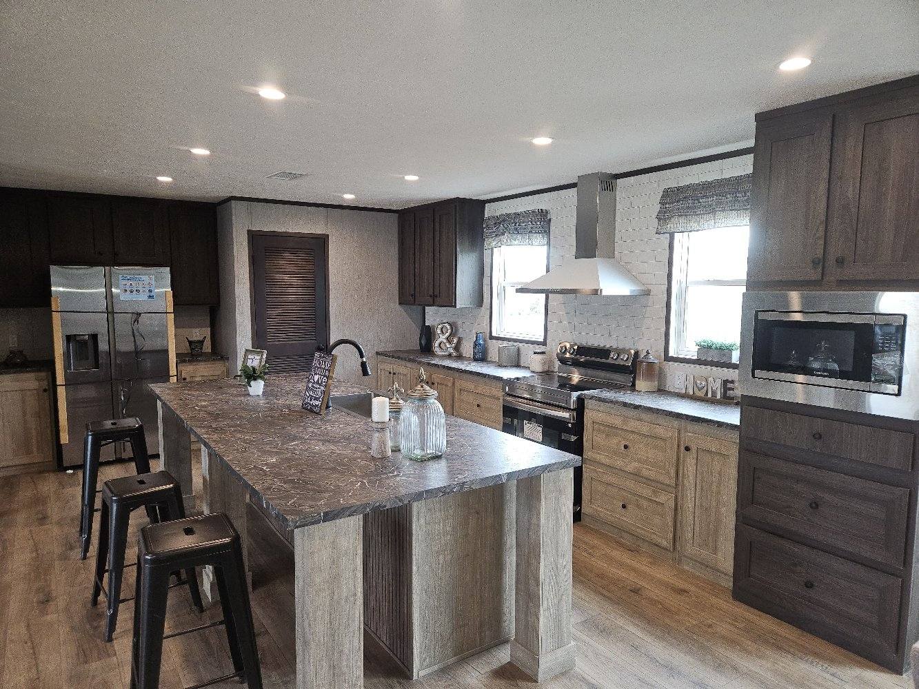 Modern kitchen with a large island and three metal stools. Dark cabinets contrast with light wooden floors and countertops. Bright, inviting ambiance.