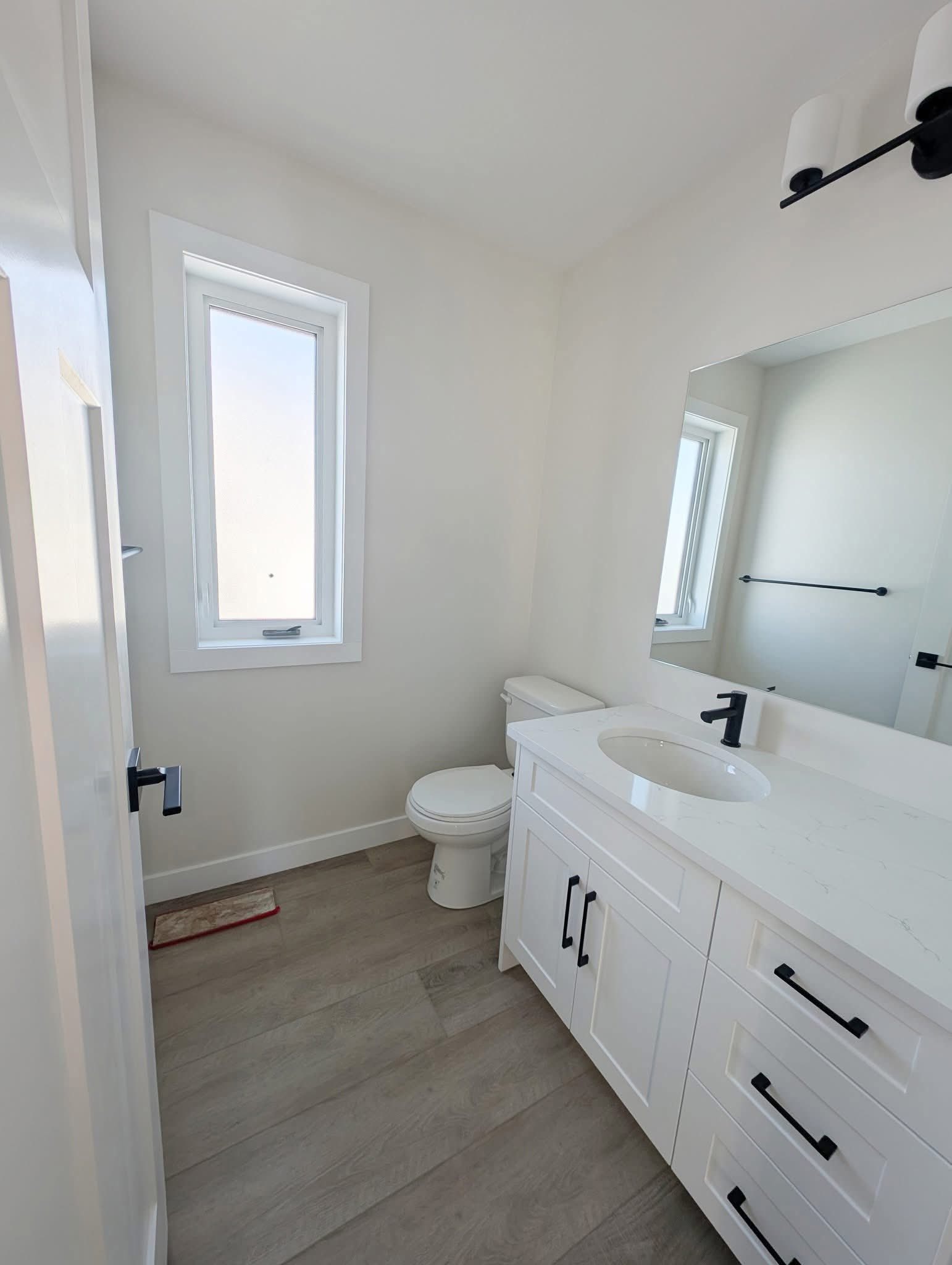 Small bathroom with a modern, minimalist design. Features a white vanity, black fixtures, a rectangular mirror, and tall window. Light wood flooring.