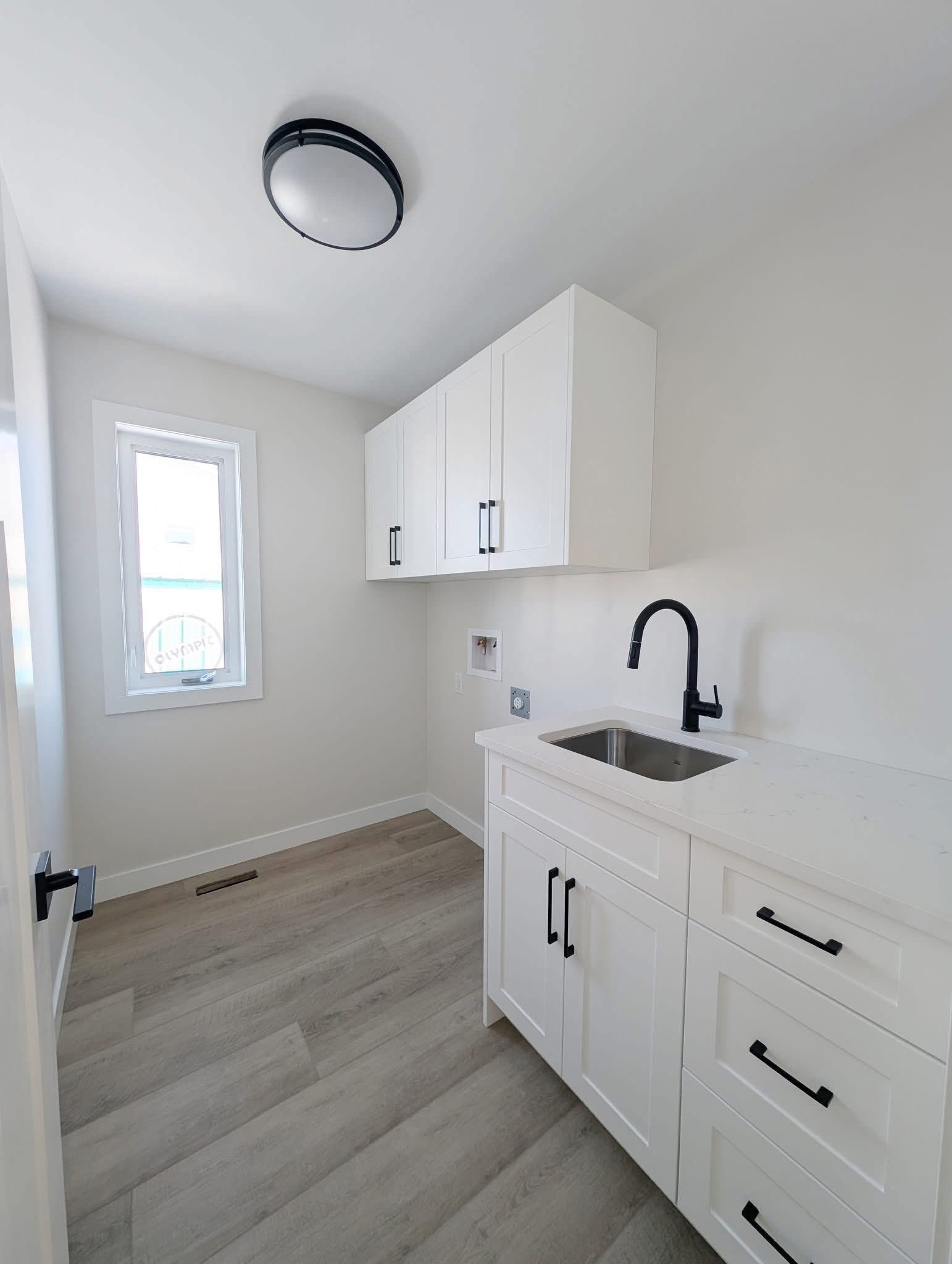 Bright laundry room with white cabinets, black handles, and a black faucet above a small sink. Light wood flooring and a circular ceiling light.