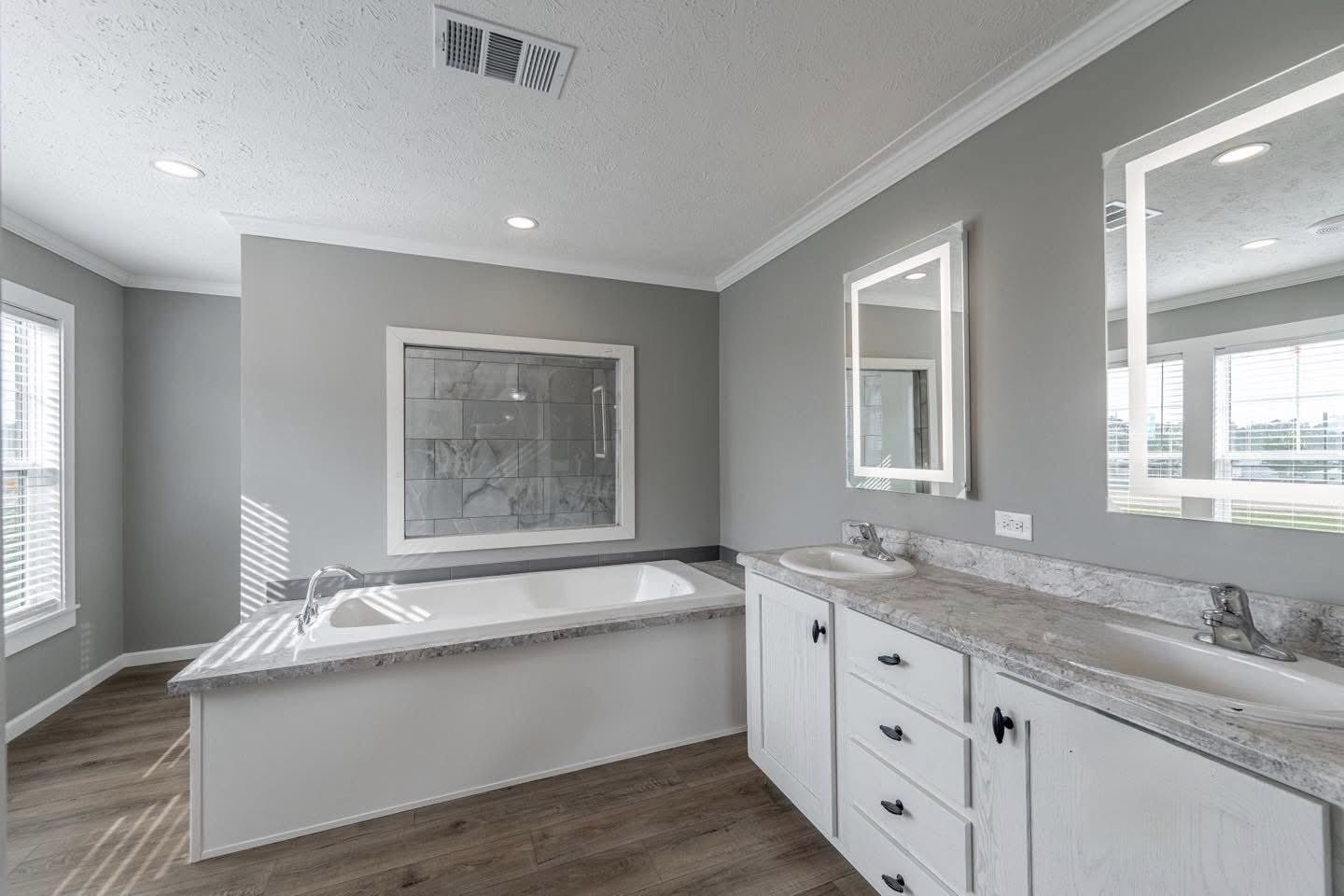 Spacious bathroom with a large tub, glass-enclosed shower, dual sinks with mirrors, and light gray walls. Natural light from multiple windows. Modern and serene.