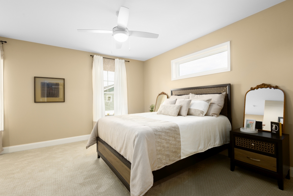 A cozy bedroom features a bed with white and beige linens, flanked by a nightstand with a mirror. Beige walls, a window, and a ceiling fan create a serene atmosphere.