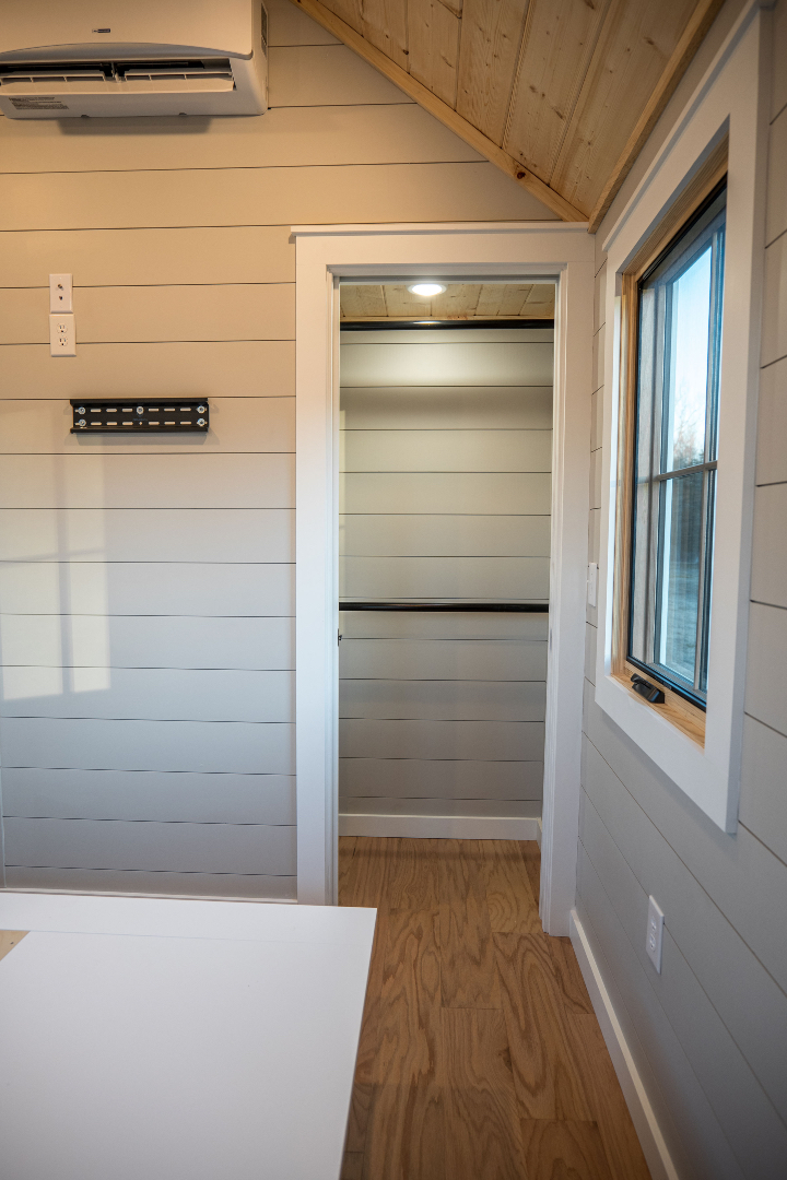 Bright interior of a small room with pale wood paneling, a window on the right, an open door revealing a closet, and a wall-mounted air conditioner above.