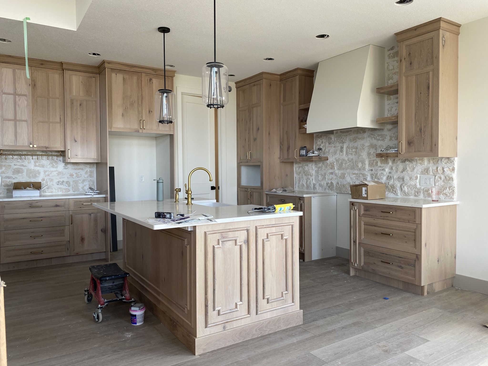 Modern kitchen under construction with light wood cabinets, stone backsplash, and a central island with white countertops. Two pendant lights hang overhead.
