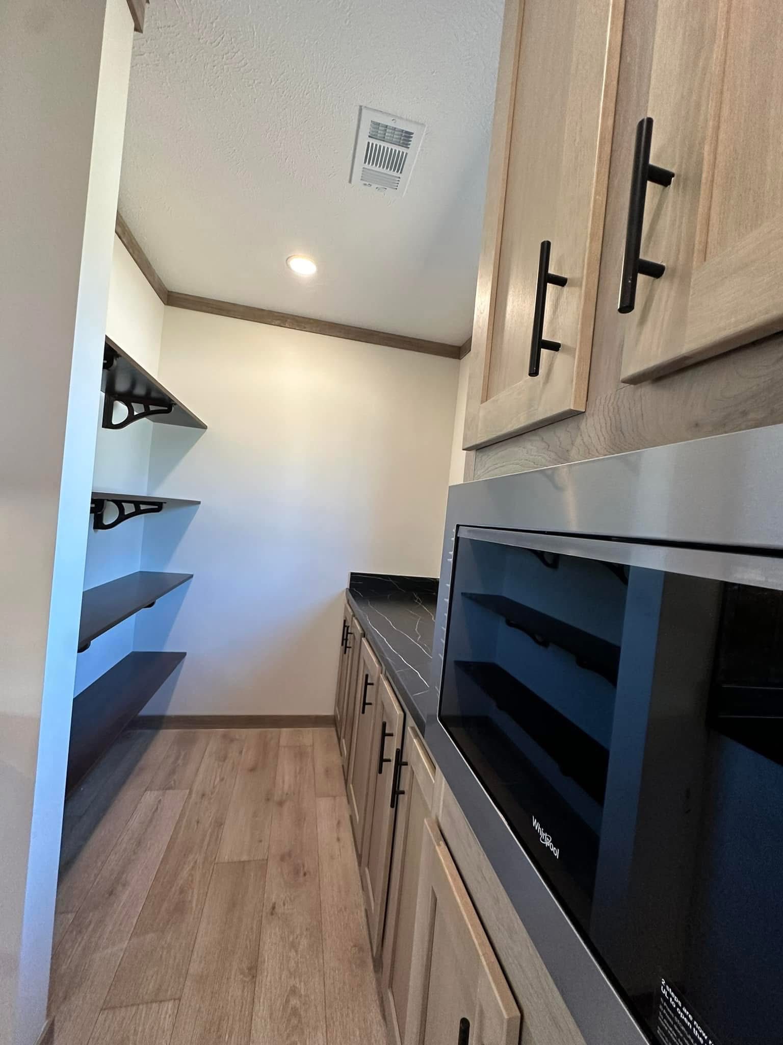 Modern pantry with wooden cabinets and black handles. Features shelves on the left and a microwave on the right, under bright overhead lighting.