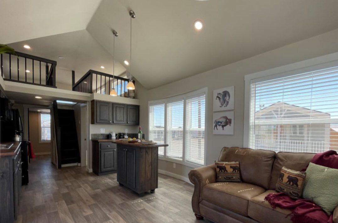 Spacious living area with high ceilings, featuring a brown couch, bear-themed pillows, and a wooden kitchen island. Bright window light adds coziness.