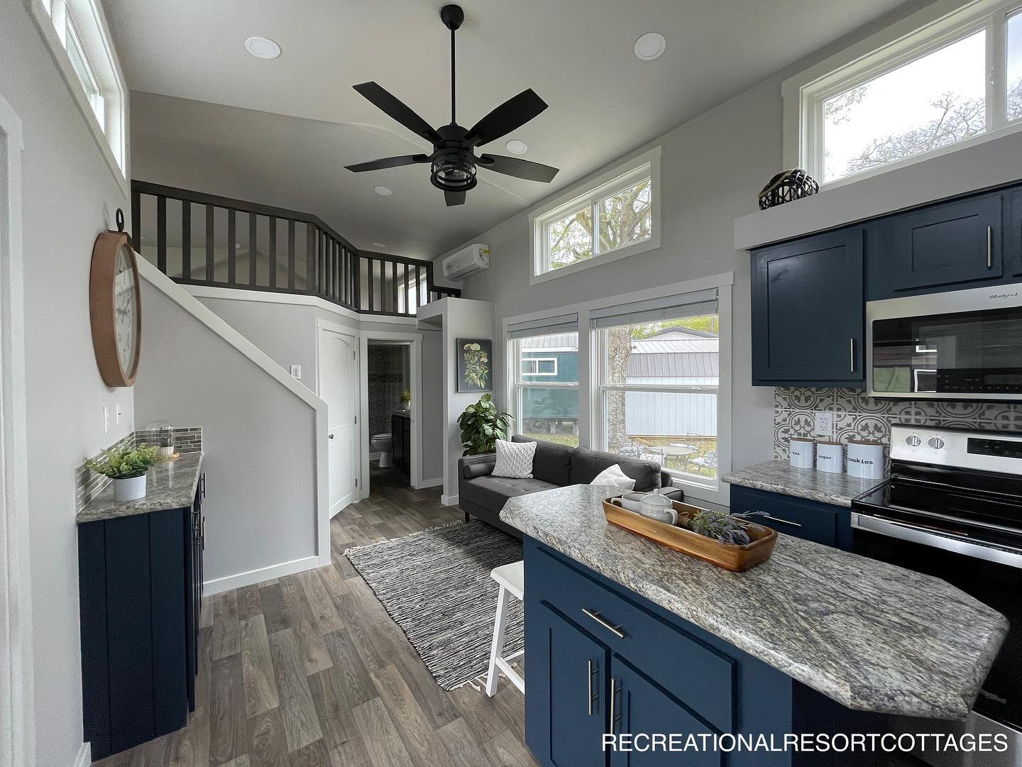 Modern tiny home interior with high ceilings, featuring a sleek kitchen with blue cabinets, granite countertops, and a living area with a gray sofa. Large windows offer ample natural light, creating an open and airy atmosphere.