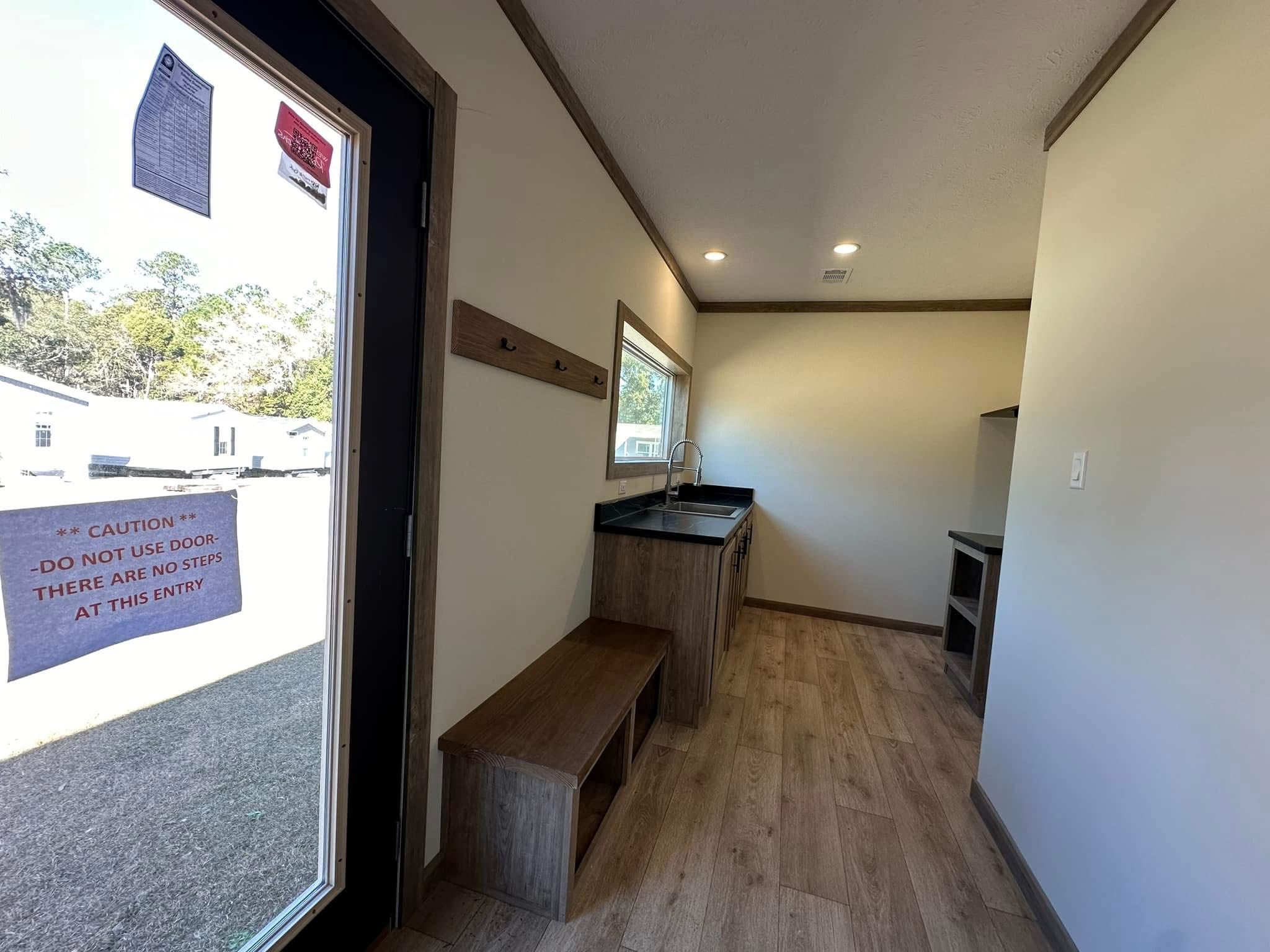 Narrow room with wooden flooring, light walls, and dark trim. Features include a bench, coat hooks, a sink under a window, and a "Caution" sign on the door.