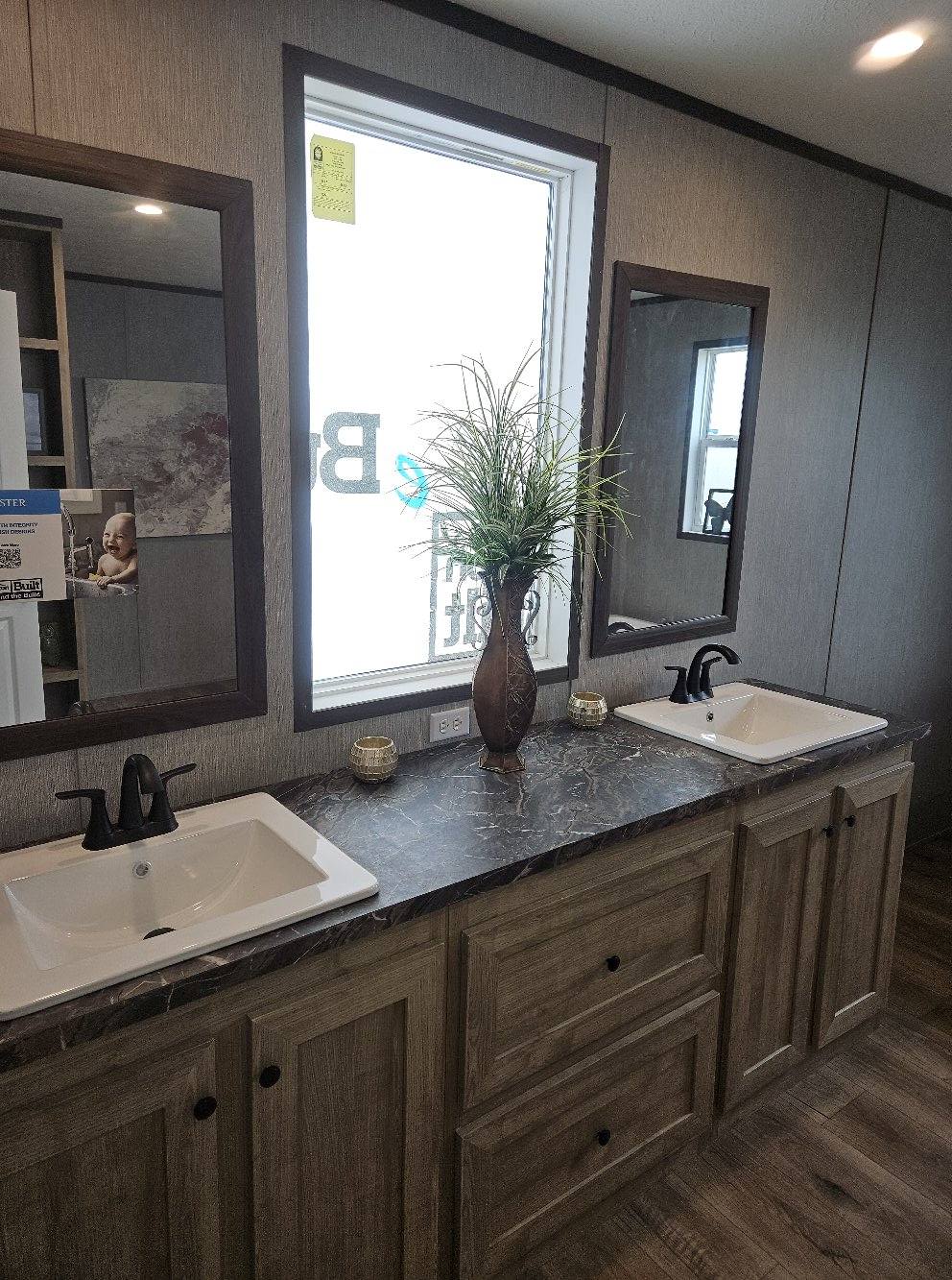 A modern bathroom with two sinks on a dark marble countertop, framed by mirrors. A vase with green plants is centered, creating a fresh, elegant tone.