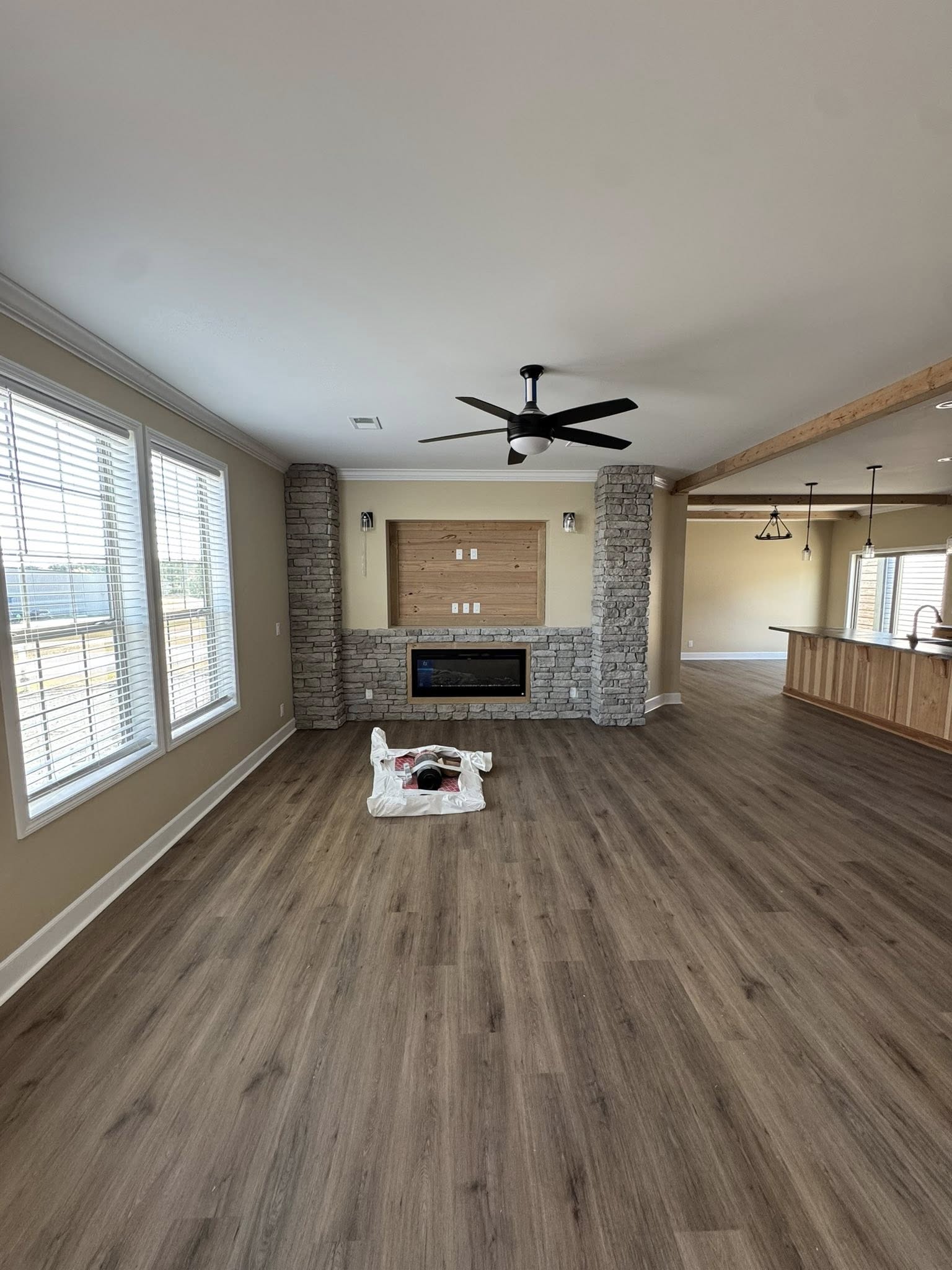 Spacious living room with wooden floor, stone fireplace, and ceiling fan. Large windows fill the space with natural light. Cozy and inviting atmosphere.
