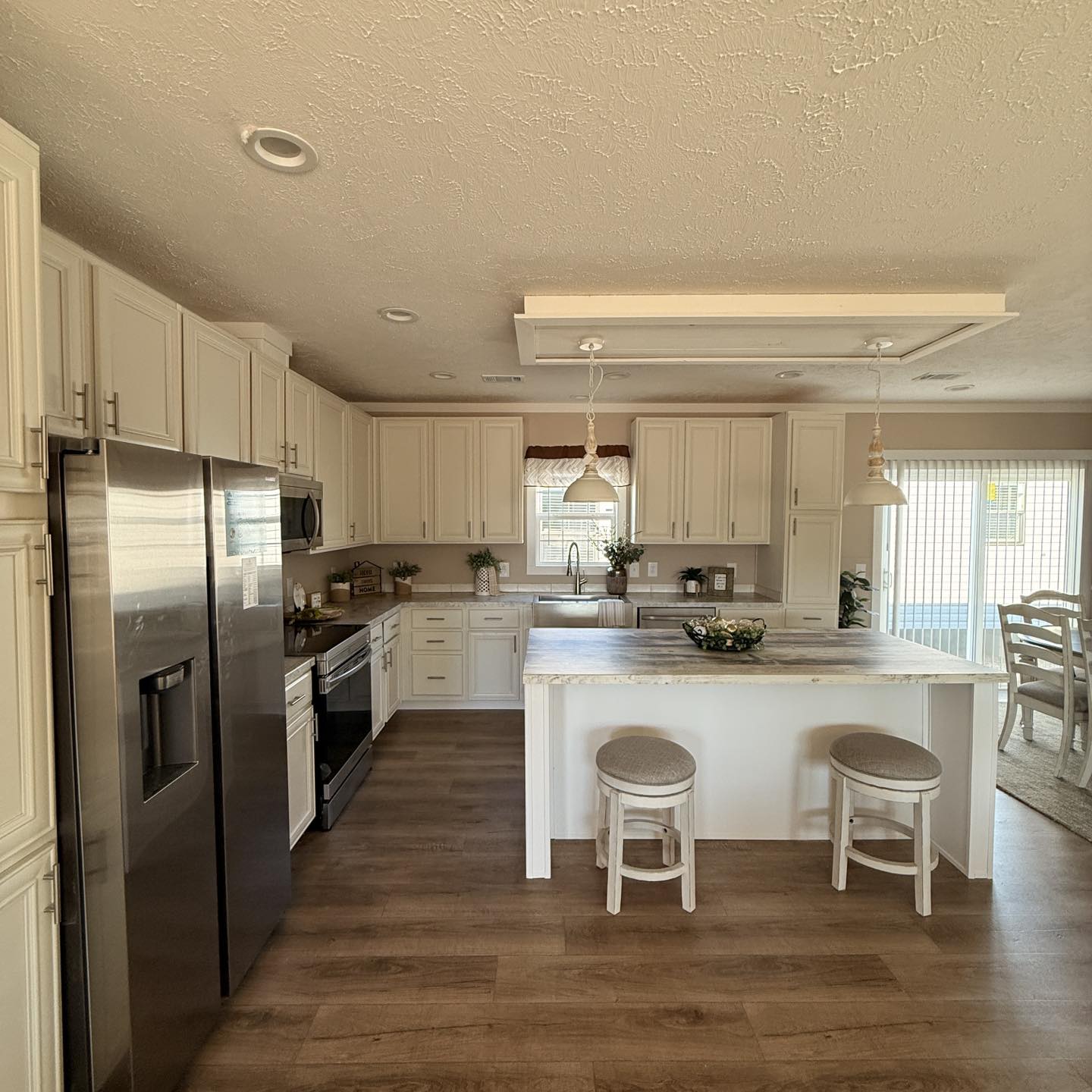 A spacious kitchen with light wooden flooring, white cabinets, and stainless steel appliances. An island with two stools and hanging lights adds warmth.