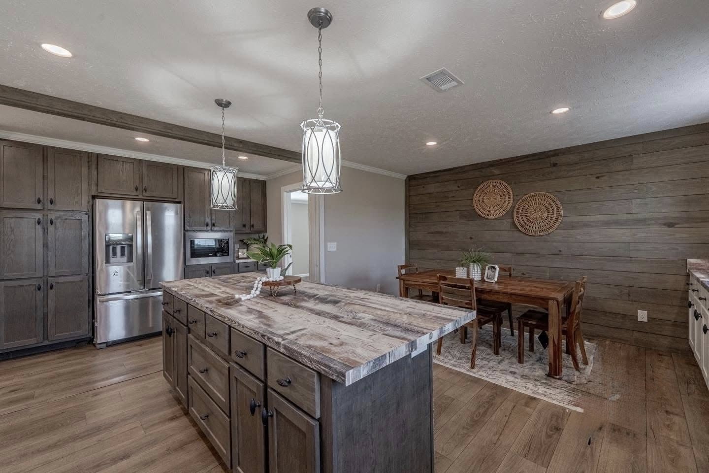 Rustic kitchen with wooden cabinets and island, pendant lights, stainless steel appliances. Dining area features wooden table with a decorative wall.