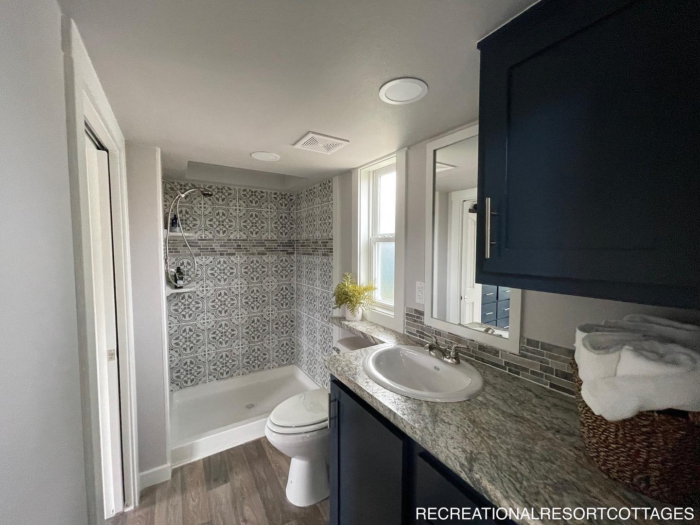 Modern bathroom with patterned tile shower, wood floor, granite countertop, and navy cabinets. Bright window light with a calming, clean vibe.